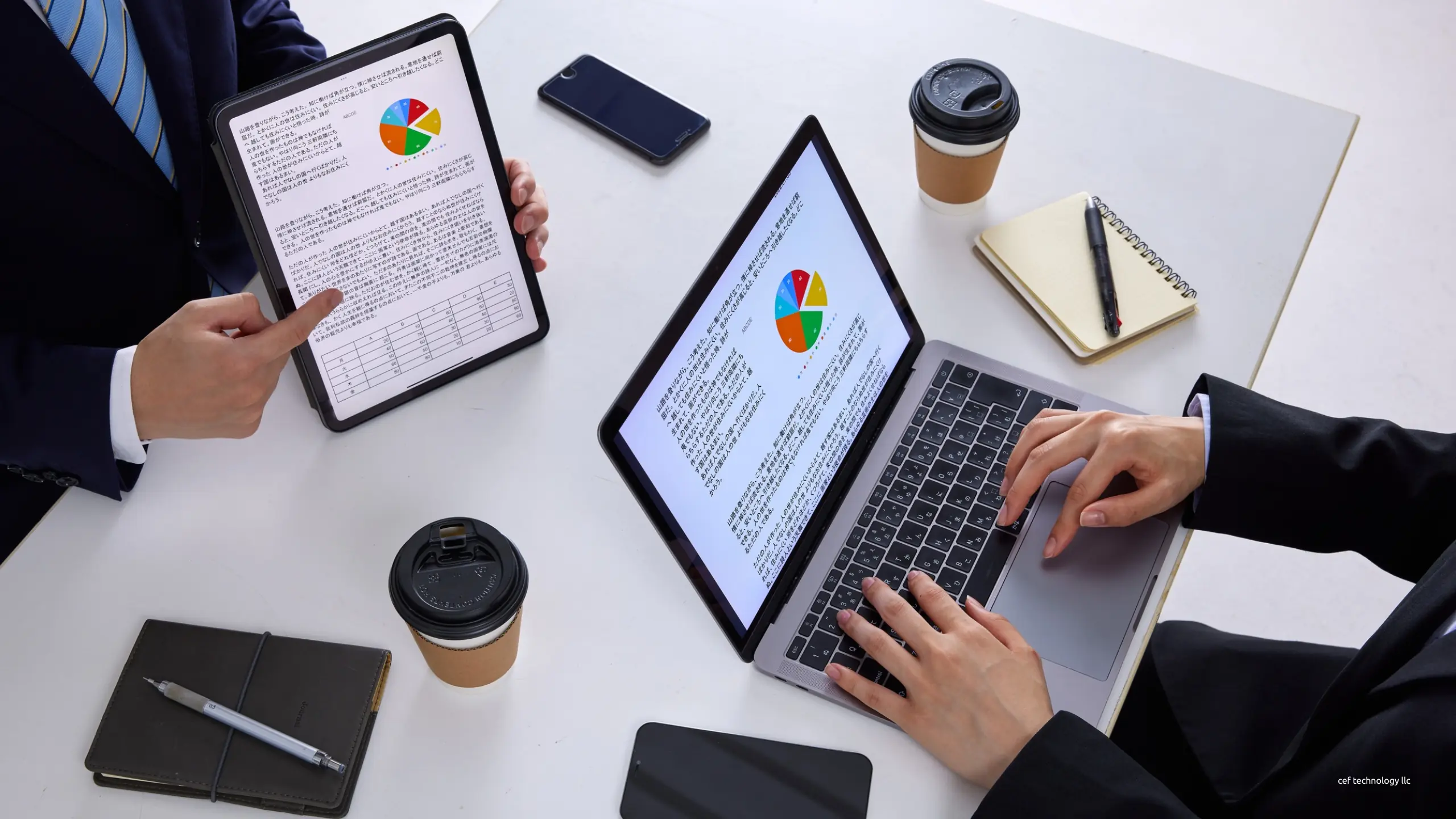 Duas pessoas em uma mesa branca analisando gráficos em um tablet e um notebook, vistas de cima.